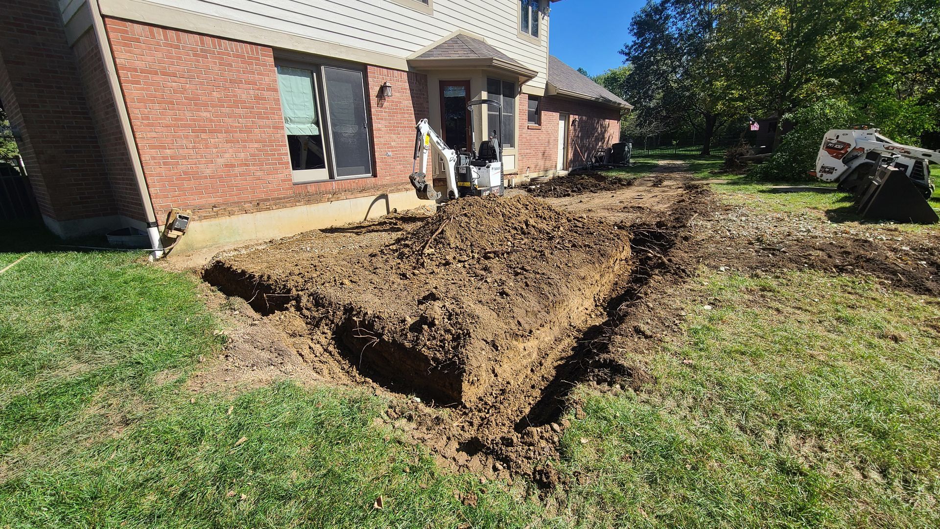 Backyard trench dug beside a brick house, with piled dirt and a man standing near the doorway