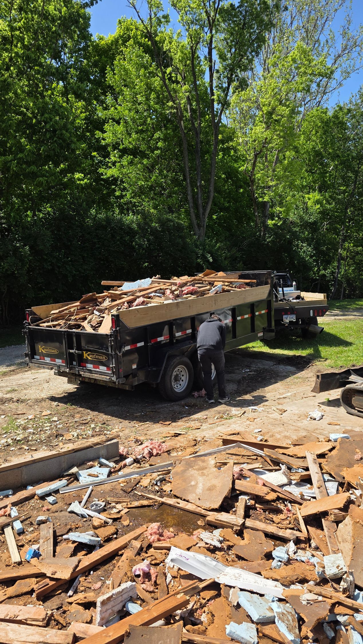 Dump trailer piled with debris at a wooded worksite, with wood scraps scattered in the foreground.