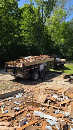 Dump trailer piled with debris at a wooded worksite, with wood scraps scattered in the foreground.
