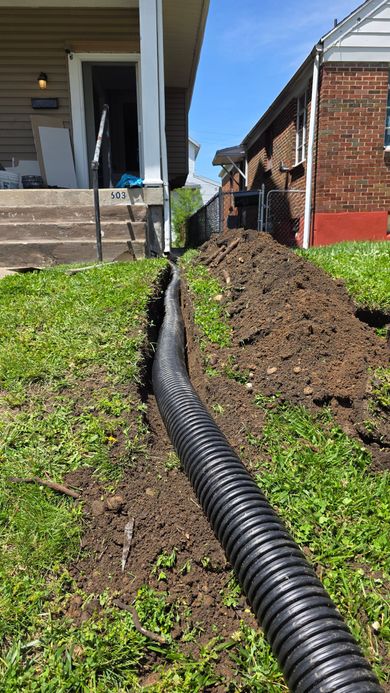 Black drainage pipe in a muddy trench between two houses, with piles of dirt along the side yard.