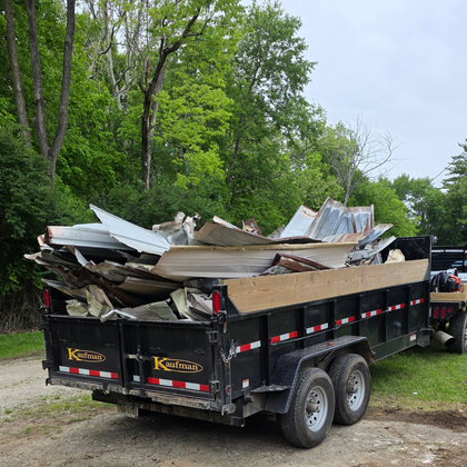 Black dump trailer loaded with scrap wood and metal on a dirt road beside trees