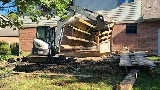Collapsed deck and stairs beside a house, with a small excavator and scattered debris in the yard.