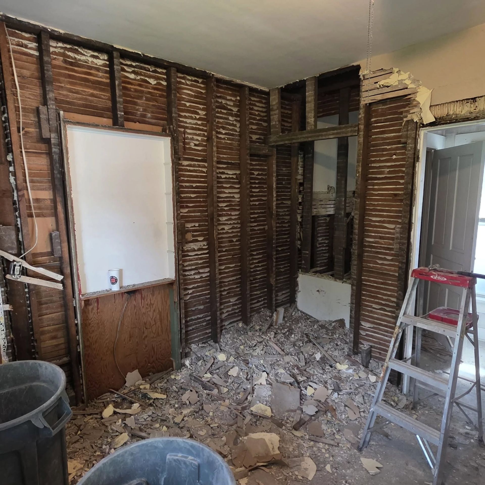 Partially demolished room with exposed wall studs, debris on the floor, and a ladder near a window