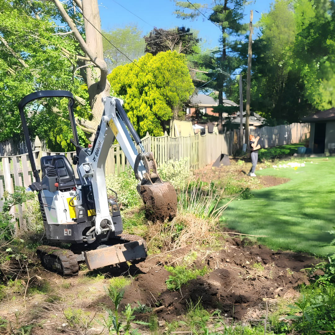 Mini excavator digging a dirt trench beside a green lawn and fence on a sunny day