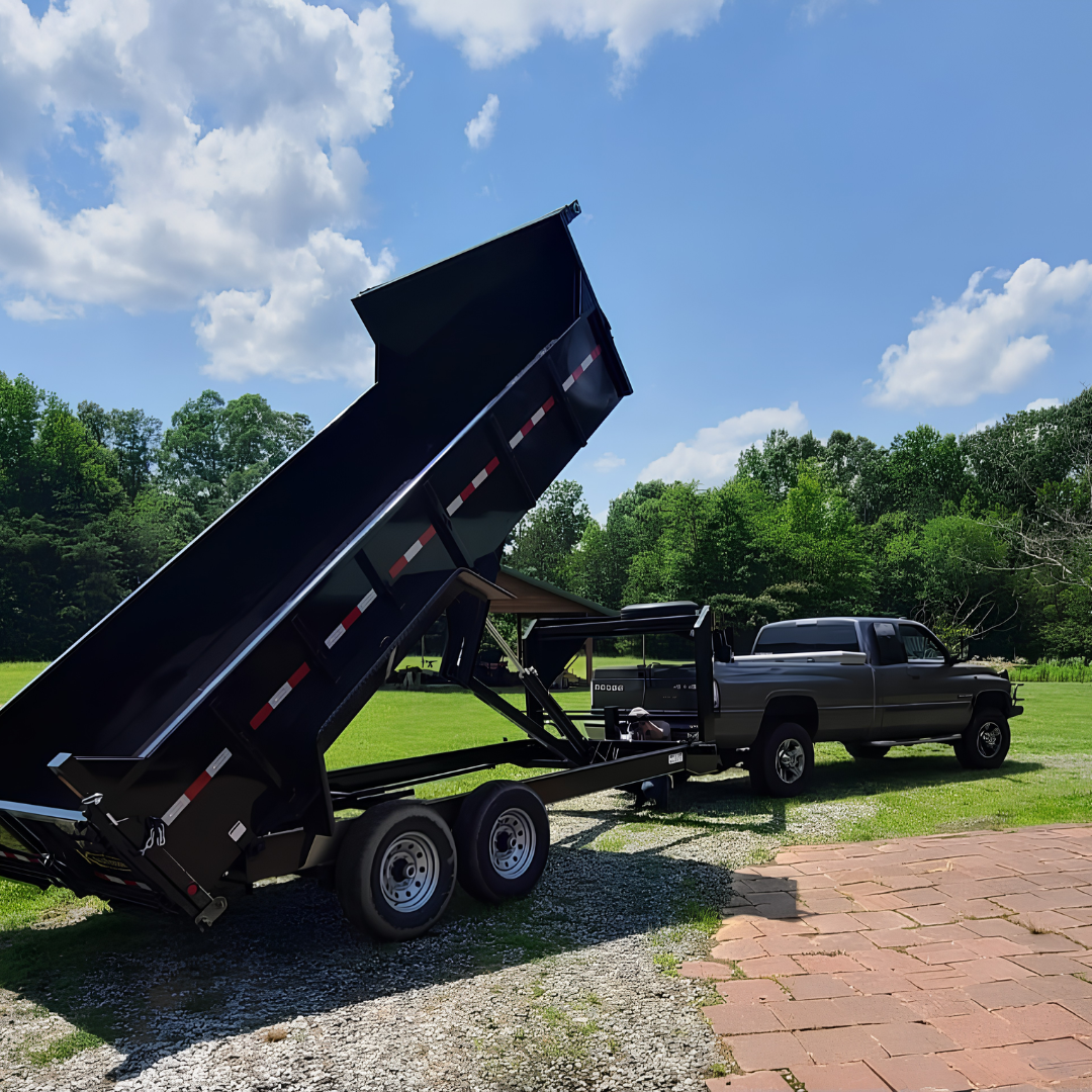Black dump trailer tipped up beside a silver pickup on a sunny gravel driveway, with trees and grass nearby