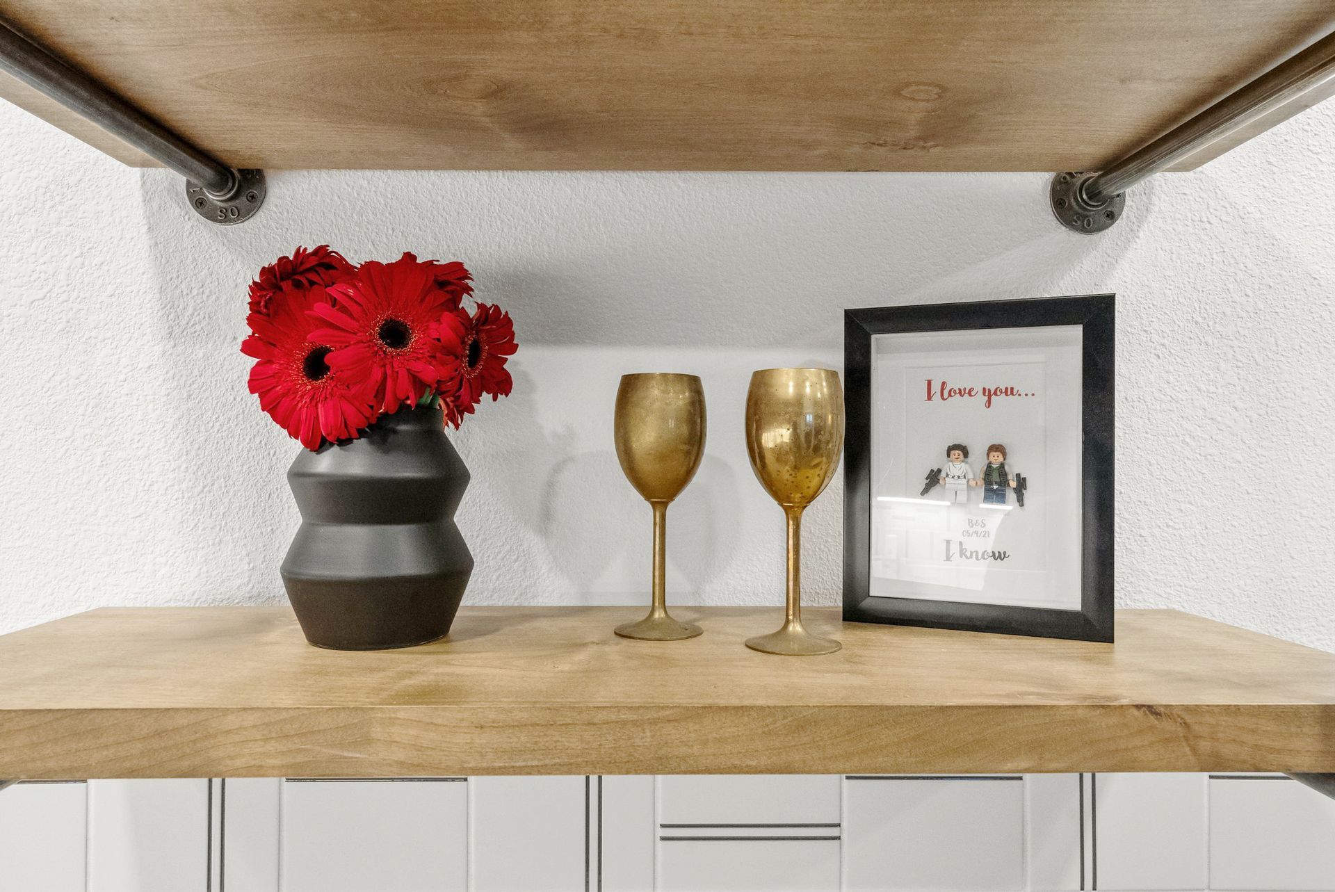 Shelf with red flowers in a black vase, two gold goblets, and a framed print on a wooden ledge.