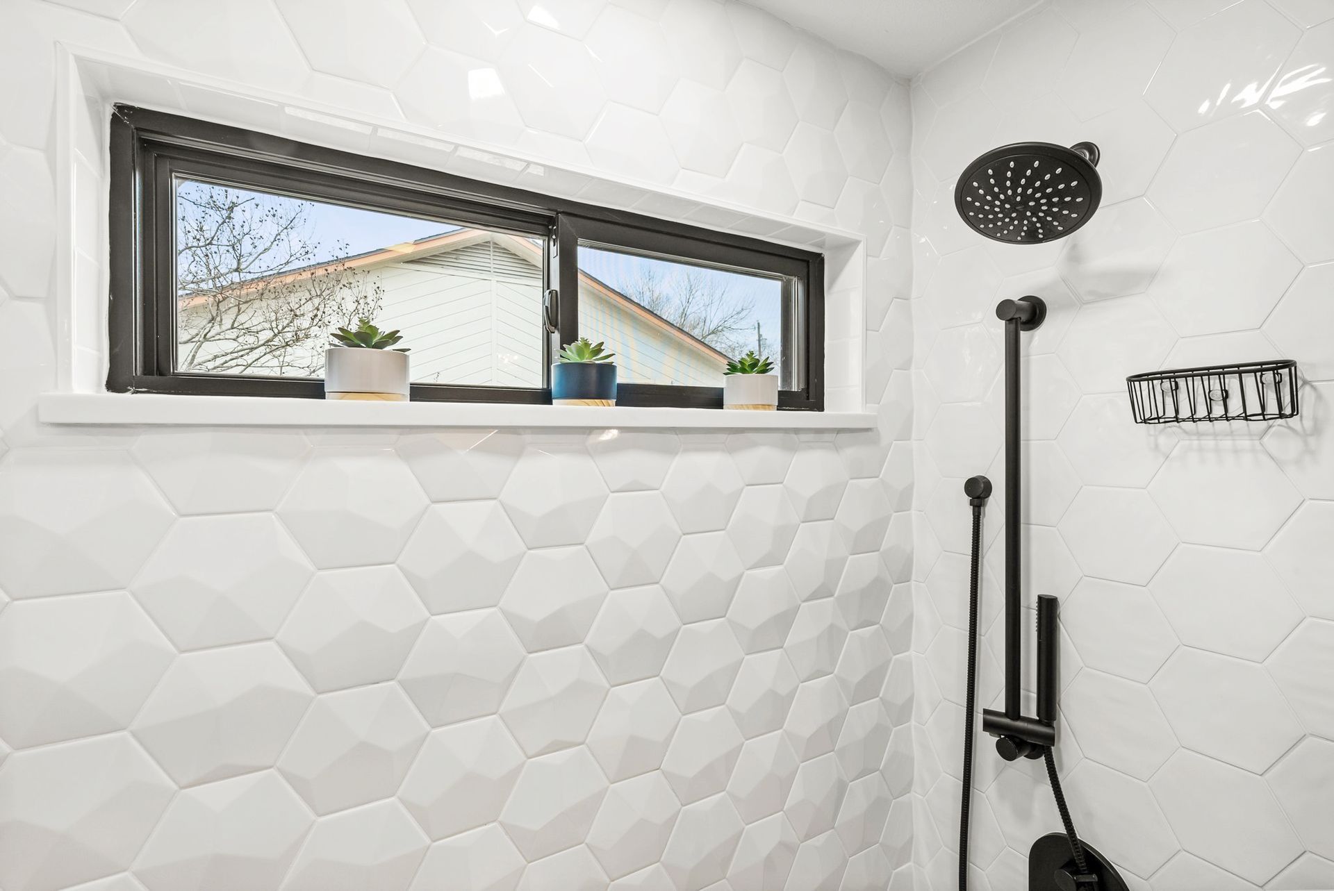 White tiled shower with hex pattern, black shower fixtures, and a small window ledge with potted plants.