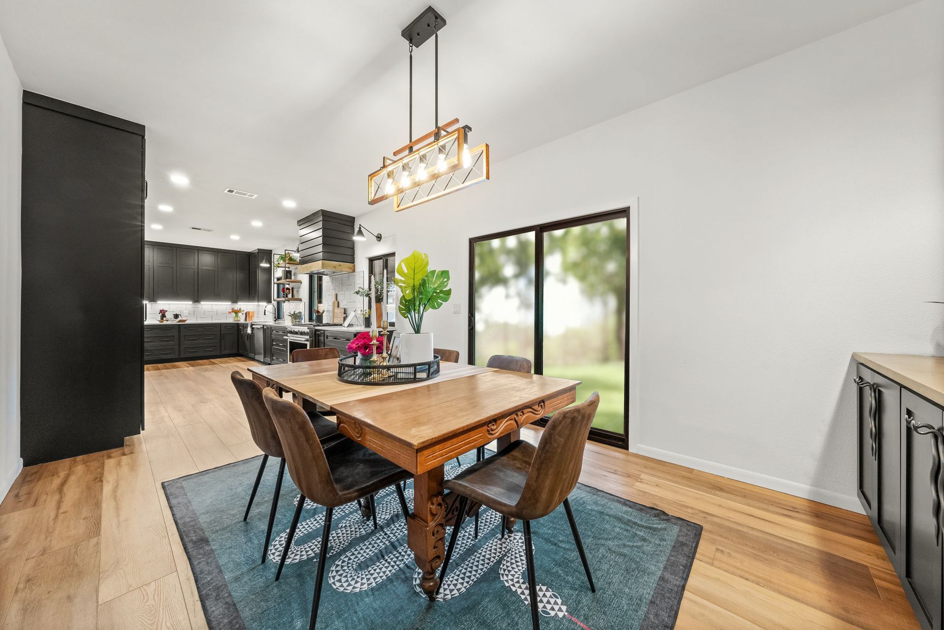 Modern dining room with wooden table, blue rug, black chairs, and large windows overlooking a backyard.