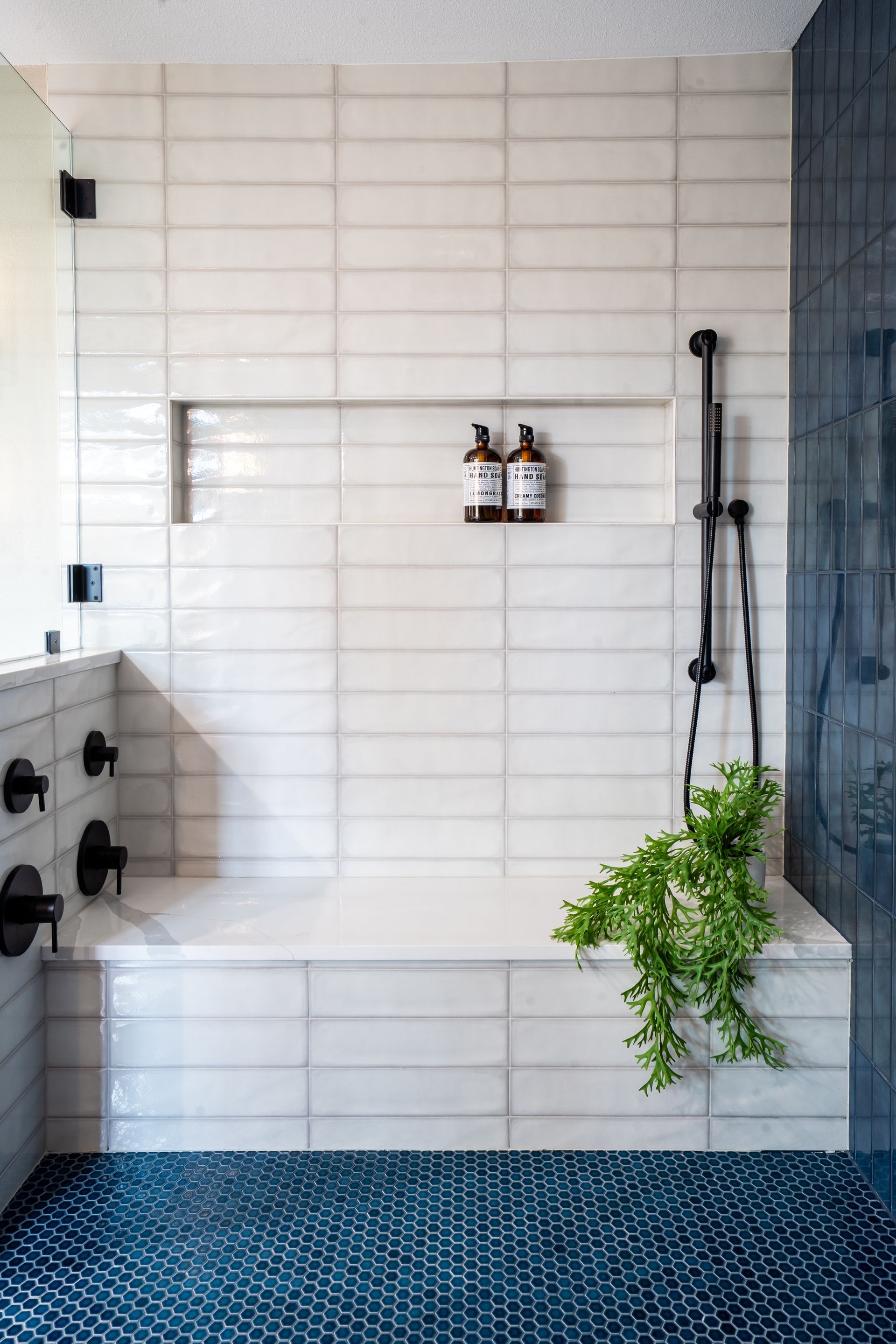 White tiled shower with built-in bench, dark blue floor, and hanging green plant.
