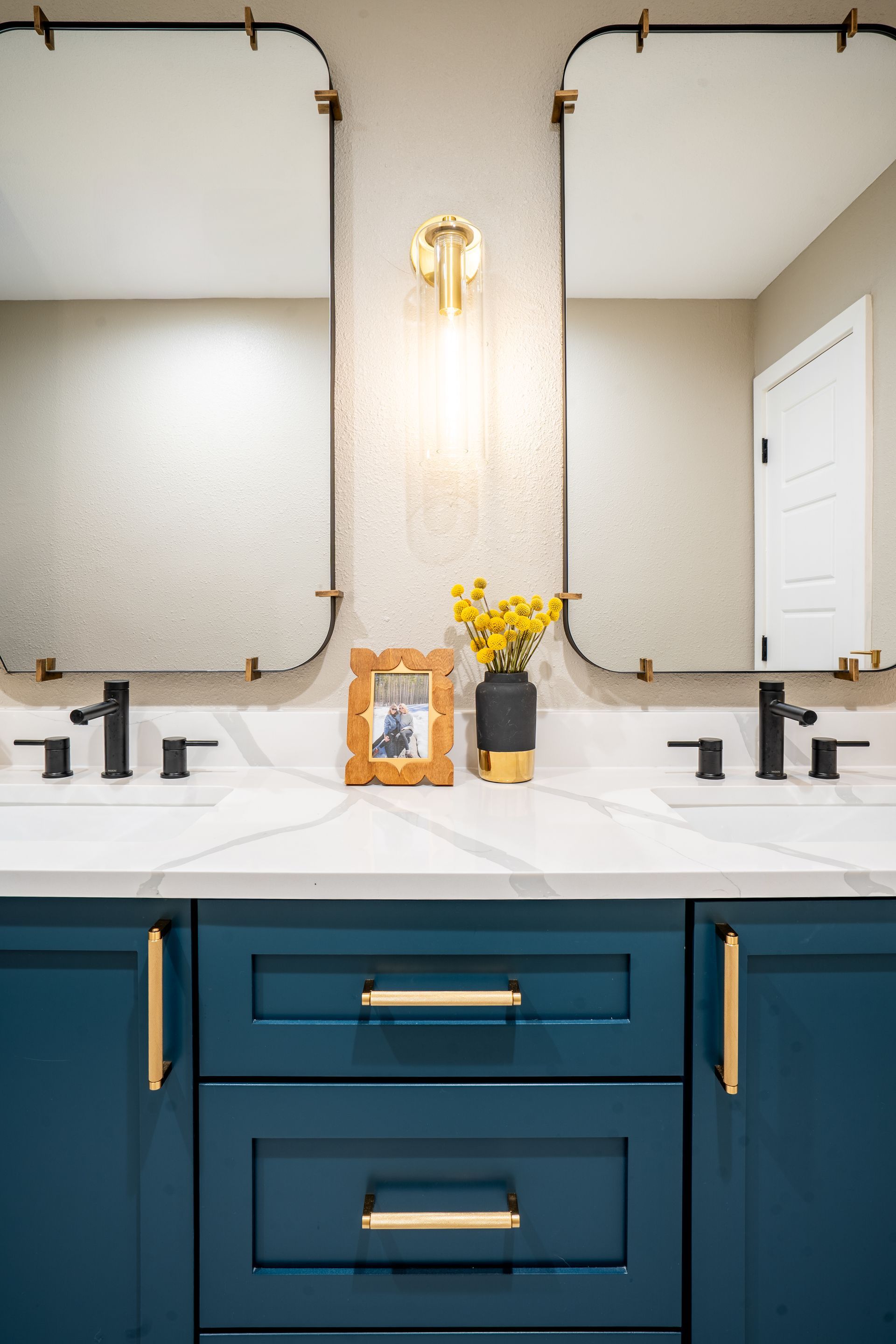 Teal bathroom vanity with double sinks, gold fixtures, mirrored wall, and flowers on the counter.