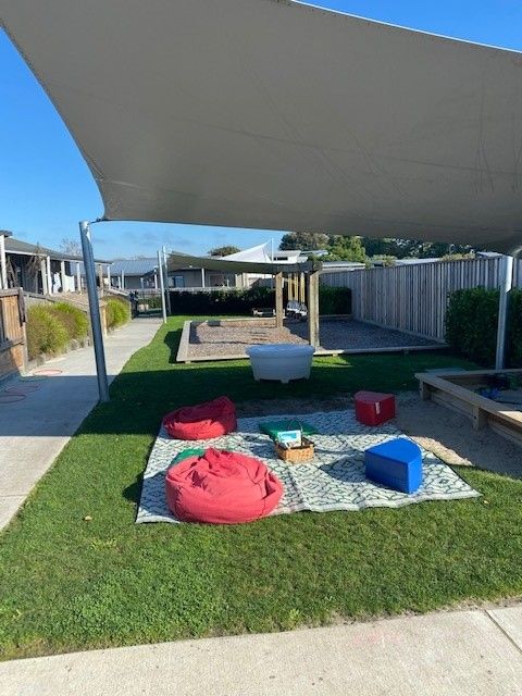 Outdoor play area with shade sail, bean bags on a mat, and sandboxes.
