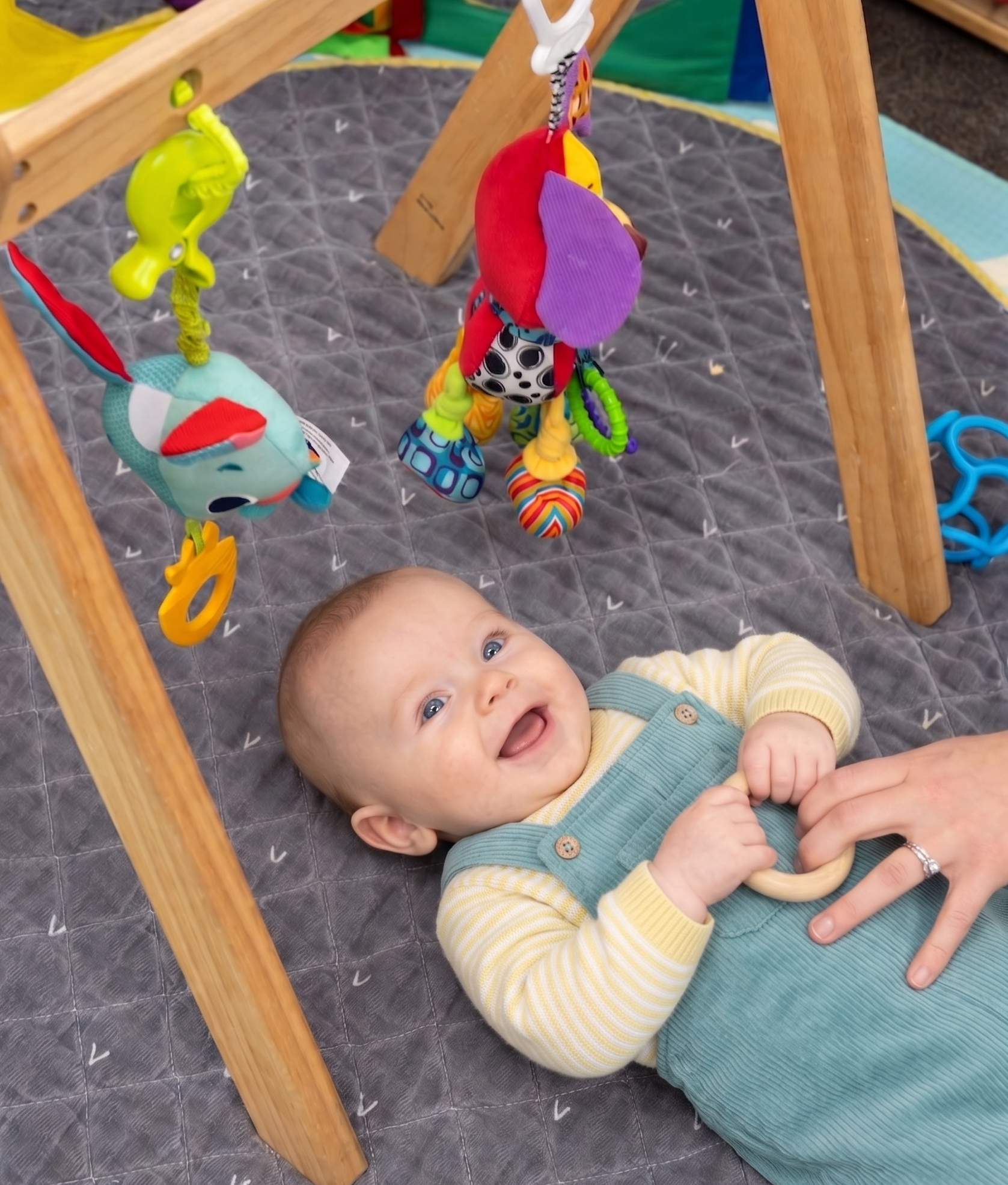 Baby laughing, playing under wooden gym with hanging toys on a gray mat.