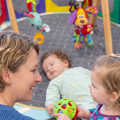 Woman and toddler looking at a toy, with sleeping baby on a playmat. Toys are overhead.