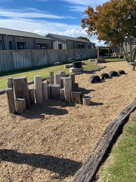 Playground with wood blocks, tires, and wood chip ground cover.  Buildings and fence in the background.