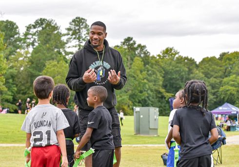 flag football Coach speaking to children outdoors on a grassy field, with trees in the background.