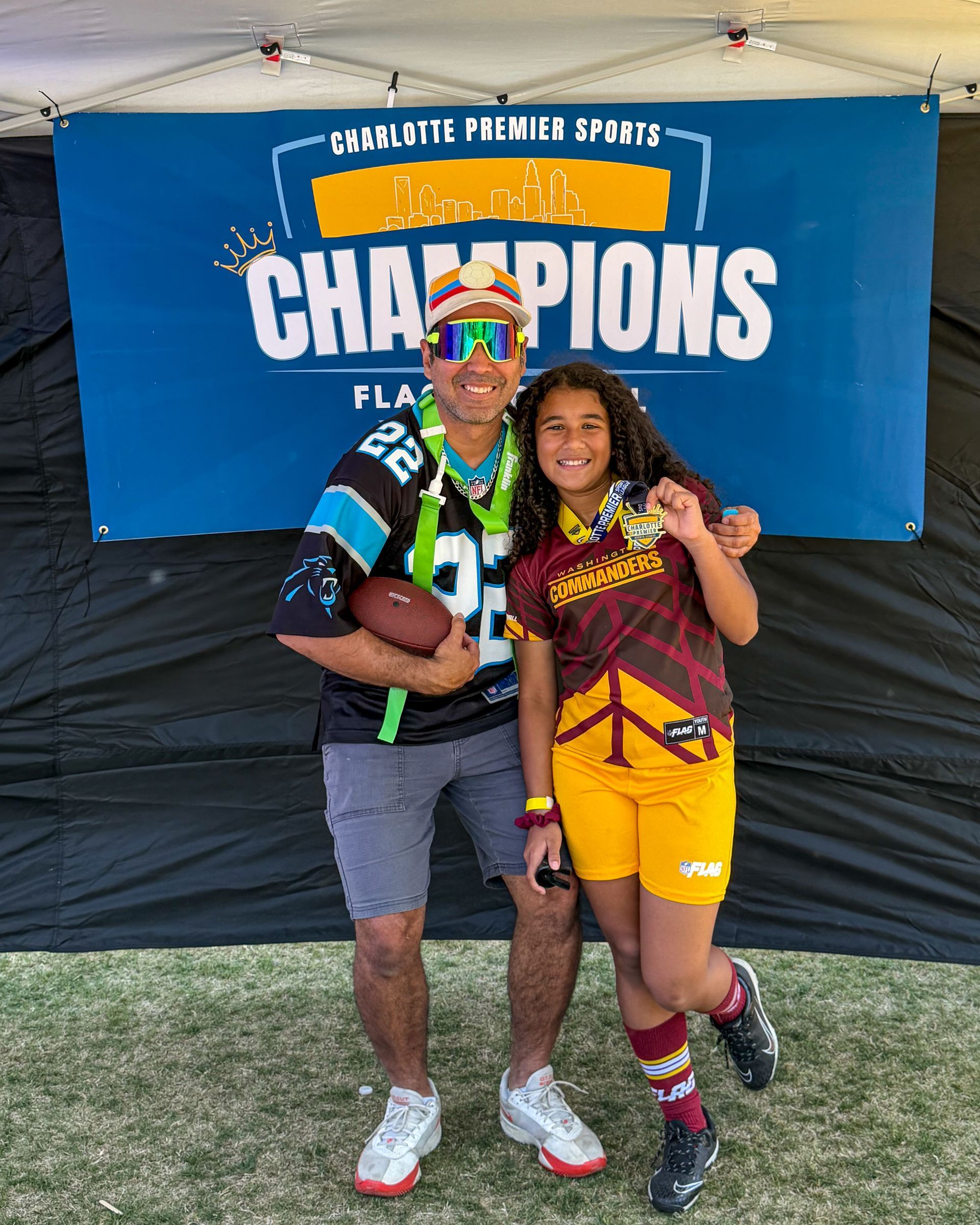 parent and child posing under a “Champions” banner at an outdoor event.
