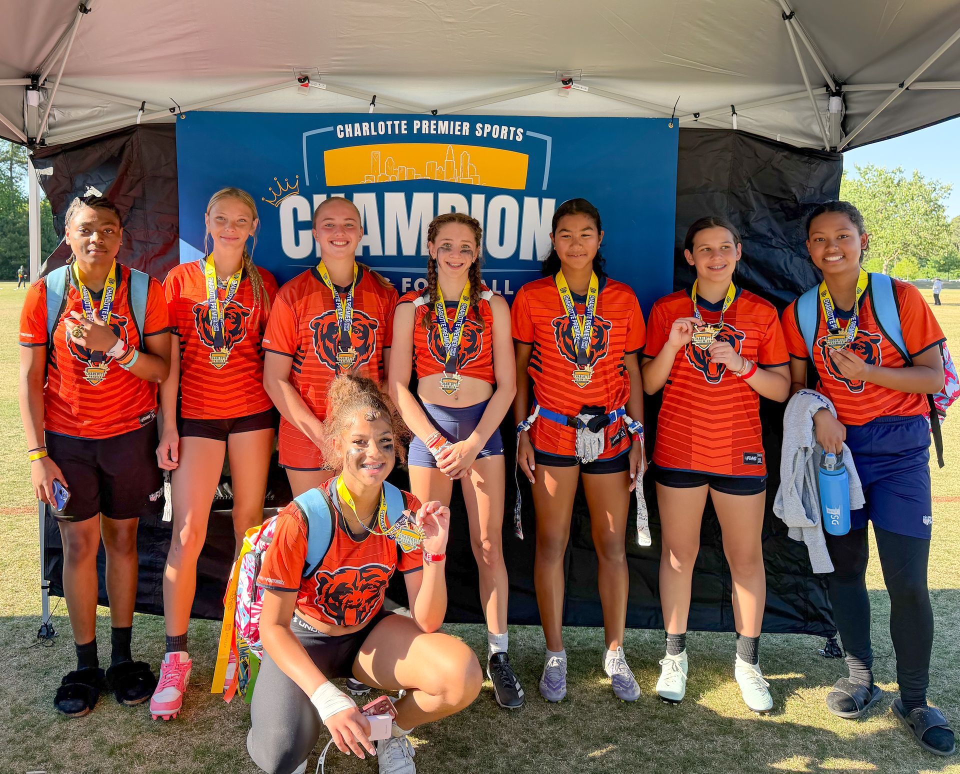 Group of flag football players in orange jerseys posing under a tent in front of a