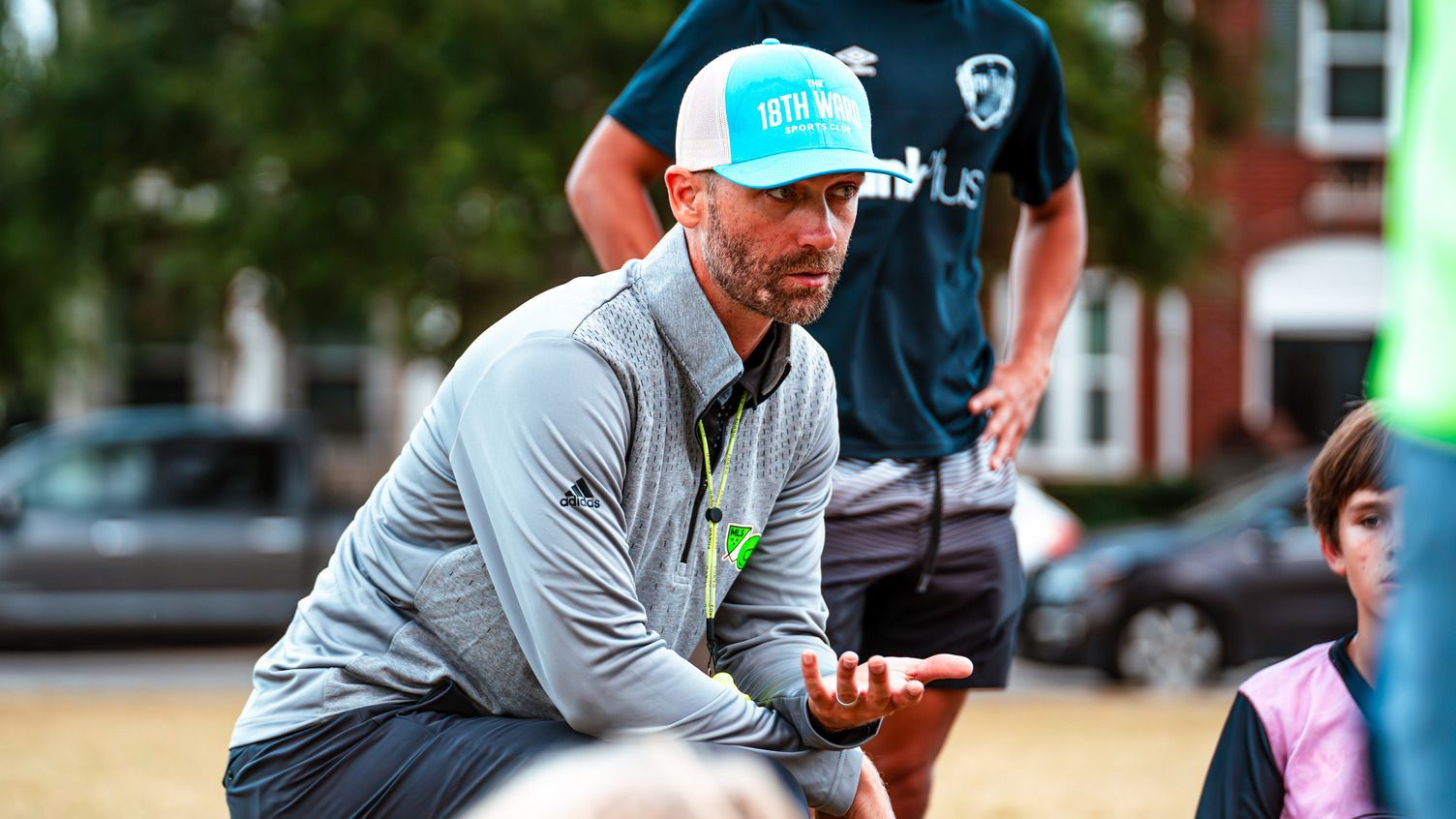 coach in a turquoise cap holds a small object outdoors, with people