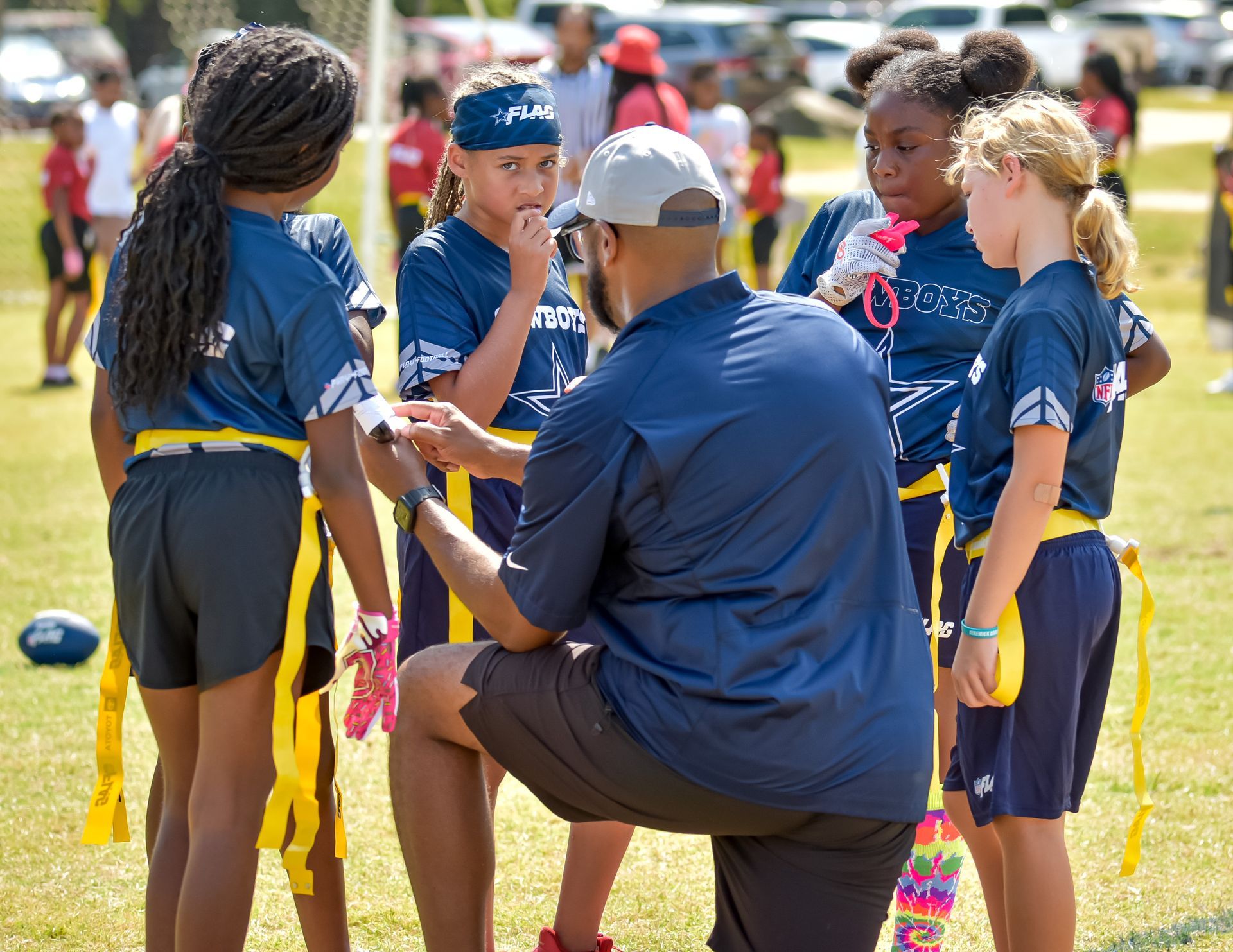 Coach kneels with youth flag football players in a grassy field huddle, wearing navy uniforms and yellow flags.