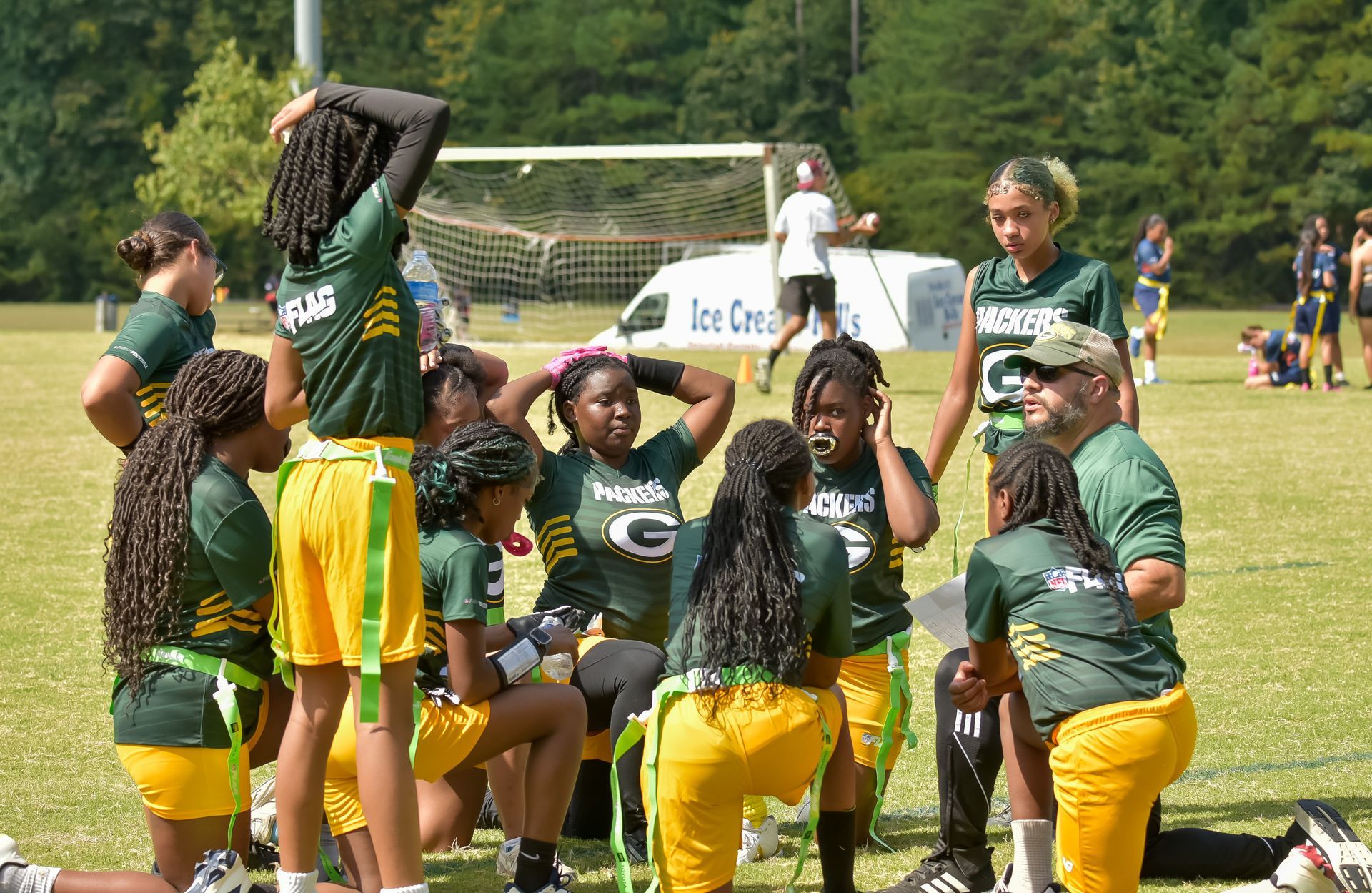 Youth flag football team huddles on a grassy field, wearing green jerseys and yellow shorts during a game break