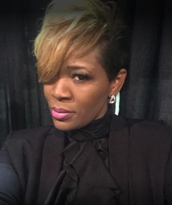 Woman with short hair, blonde bangs, pink lipstick, wearing a black blazer.