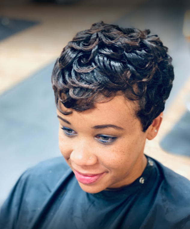 Woman with a short, curly hairstyle, wearing a blue salon cape and makeup, in a salon.