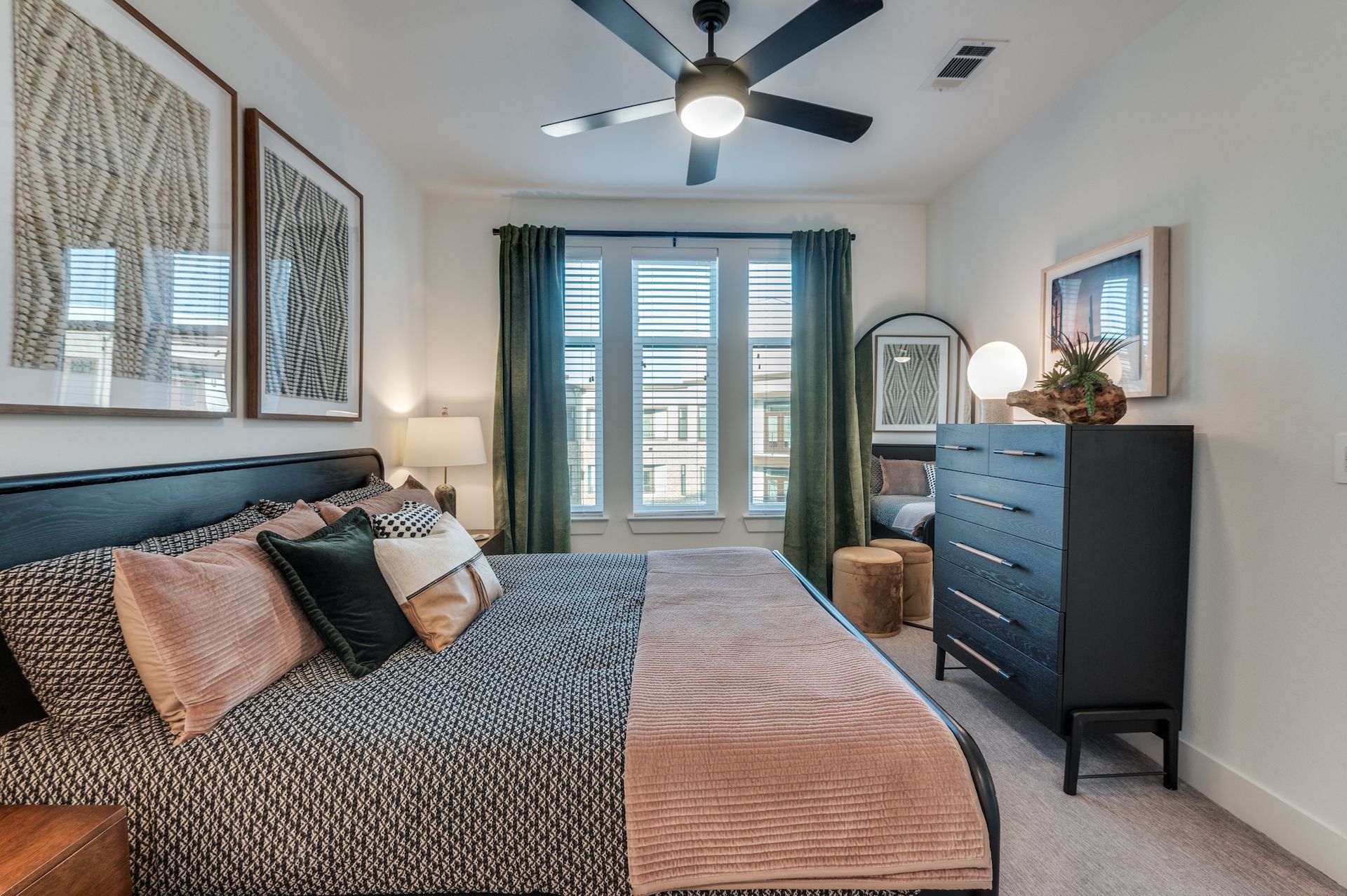 A modern bedroom with a black headboard, patterned bedding, dark dresser, and olive green curtains over a large window.