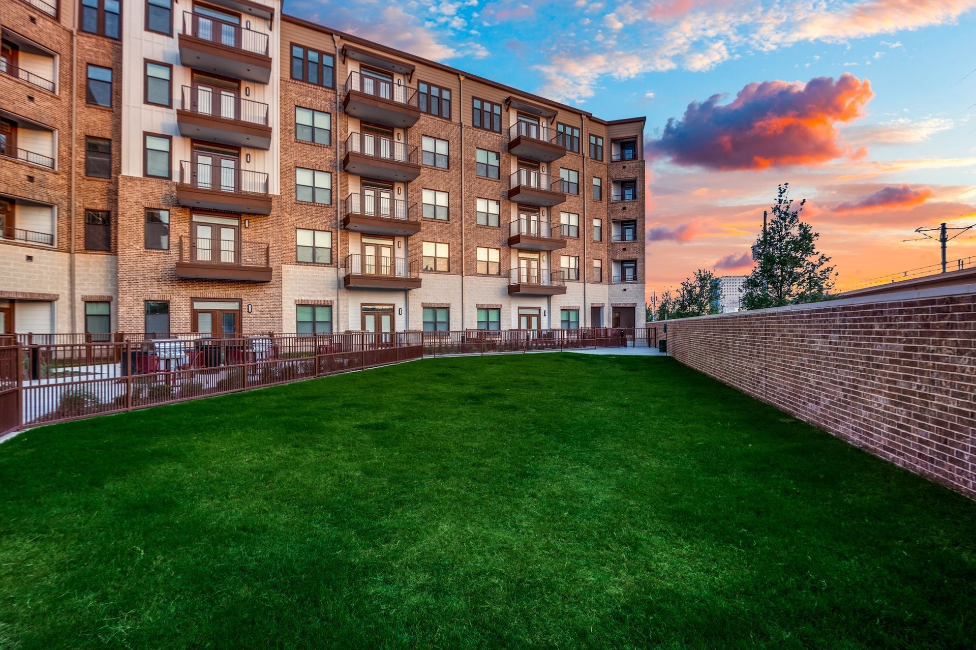 A grassy courtyard in front of a multi-story brick apartment building under a sunset sky.