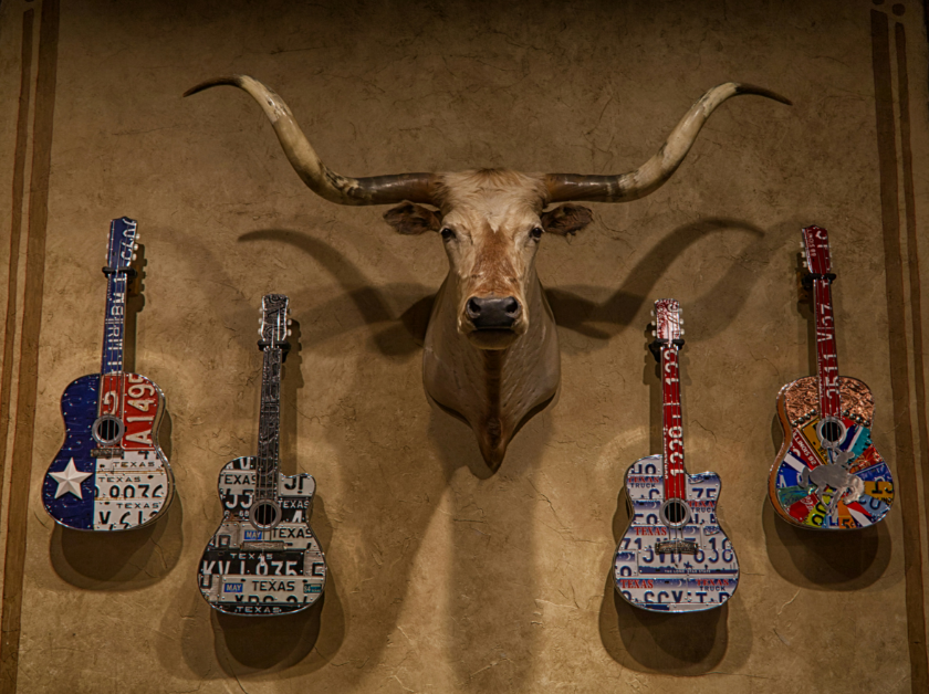 A mounted longhorn skull above five guitars decorated with license plates, on a brown wall.