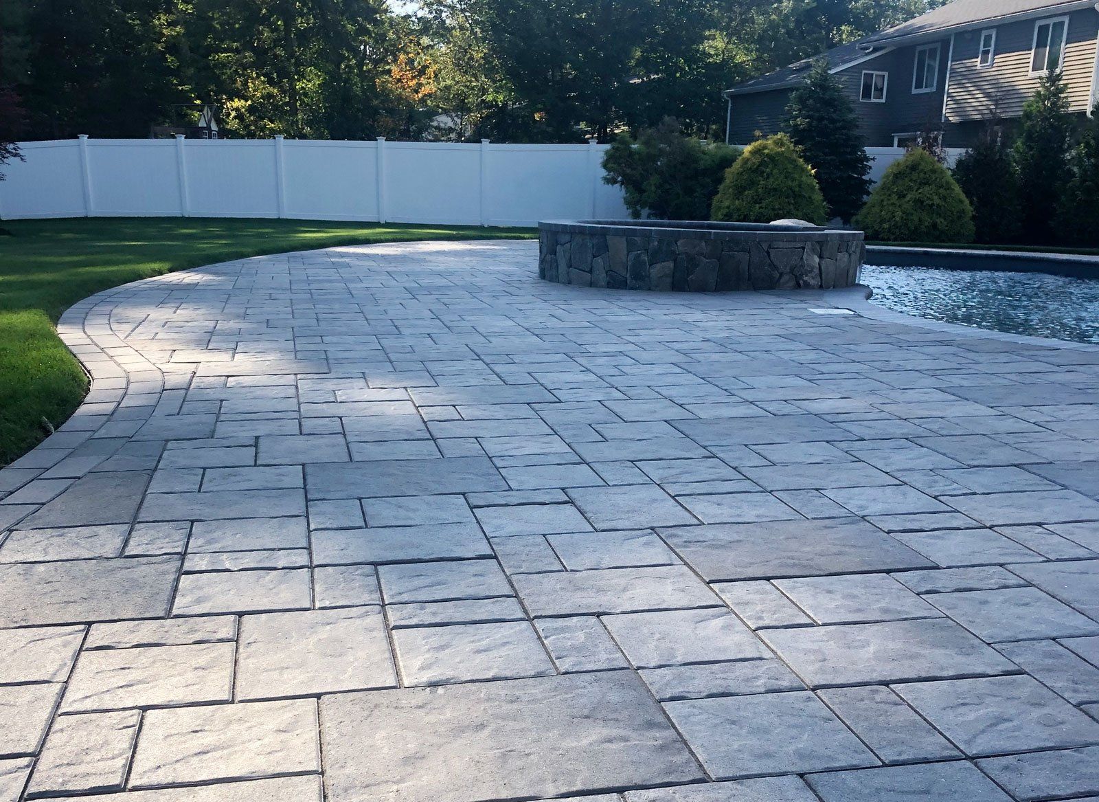 Stone patio with fire pit, bordering a pool, green lawn, and white fence.