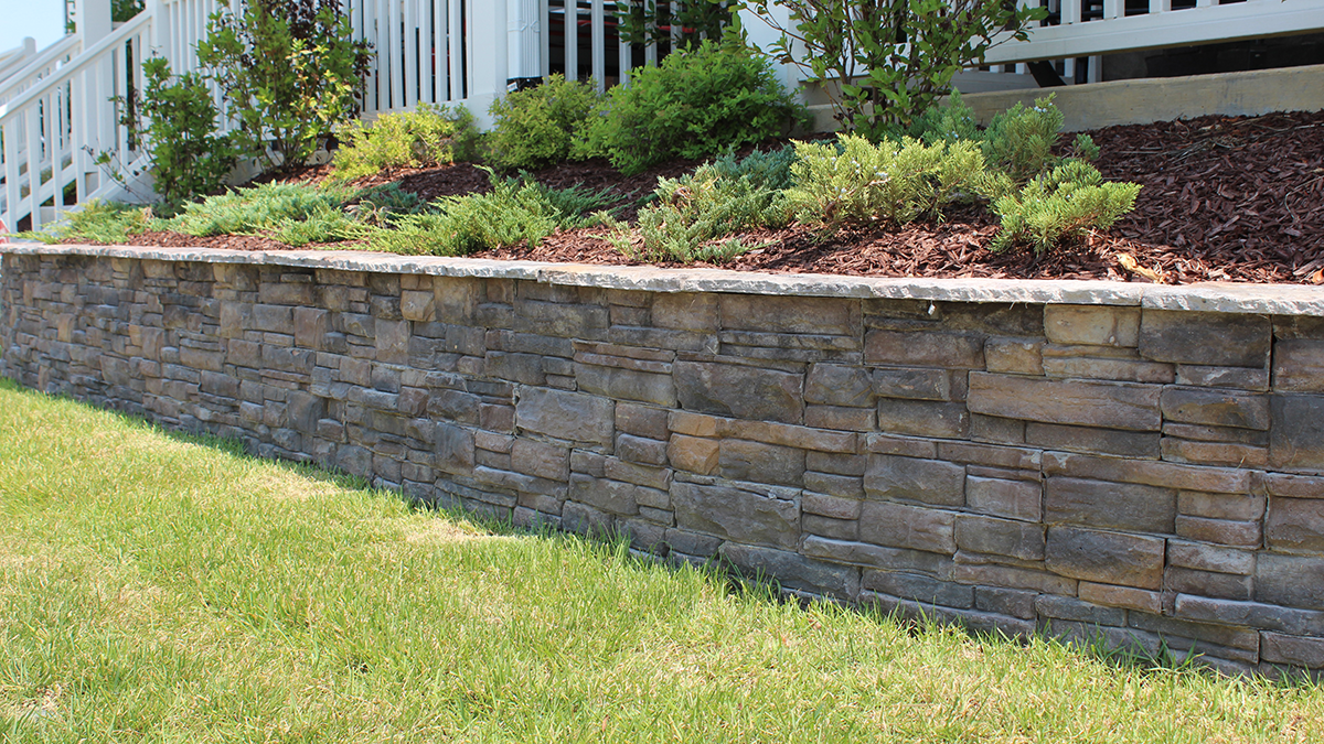 Stone retaining wall with greenery and mulch, lawn in the foreground, and a white house in the background.