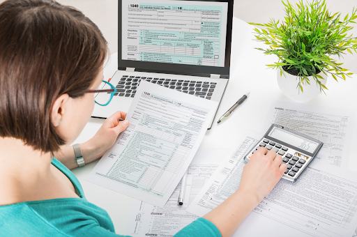 Woman using a laptop and calculator, preparing taxes with forms and documents on a white desk.