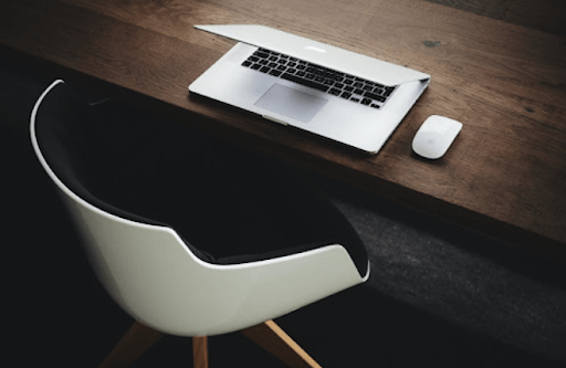Laptop and mouse on a dark wooden desk, near a modern white and black chair.