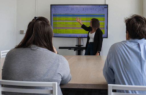 Woman presenting data on screen to two people seated at a table.