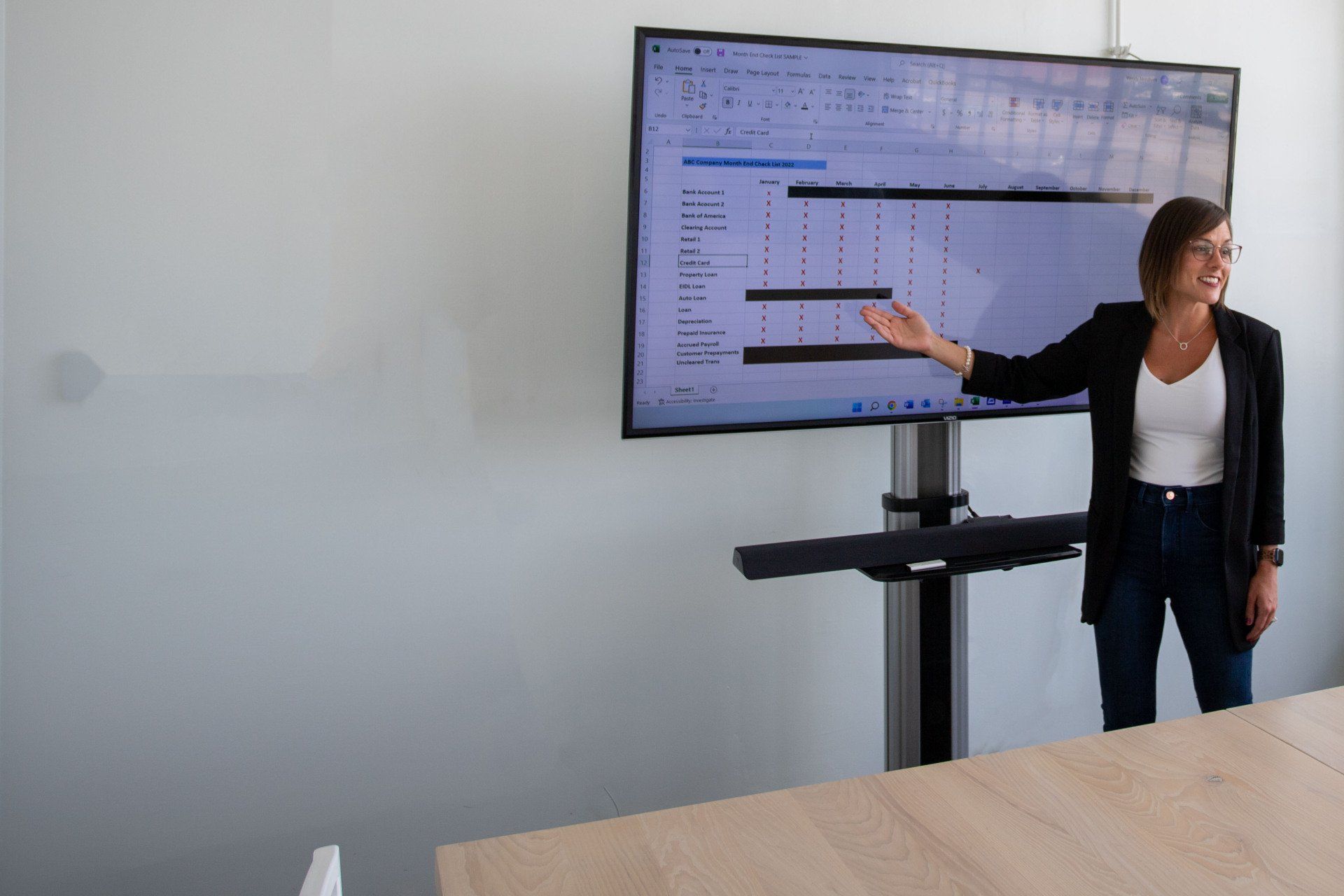 Woman presenting at a screen displaying a spreadsheet, in a bright room with a wooden table.