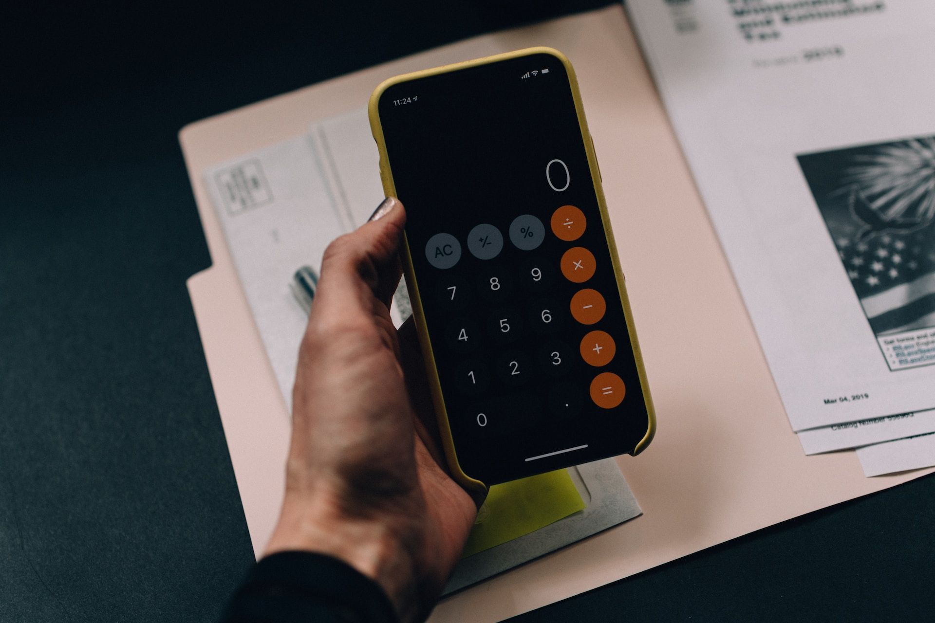 Hand holding phone displaying a calculator; documents and pen are nearby on a desk.