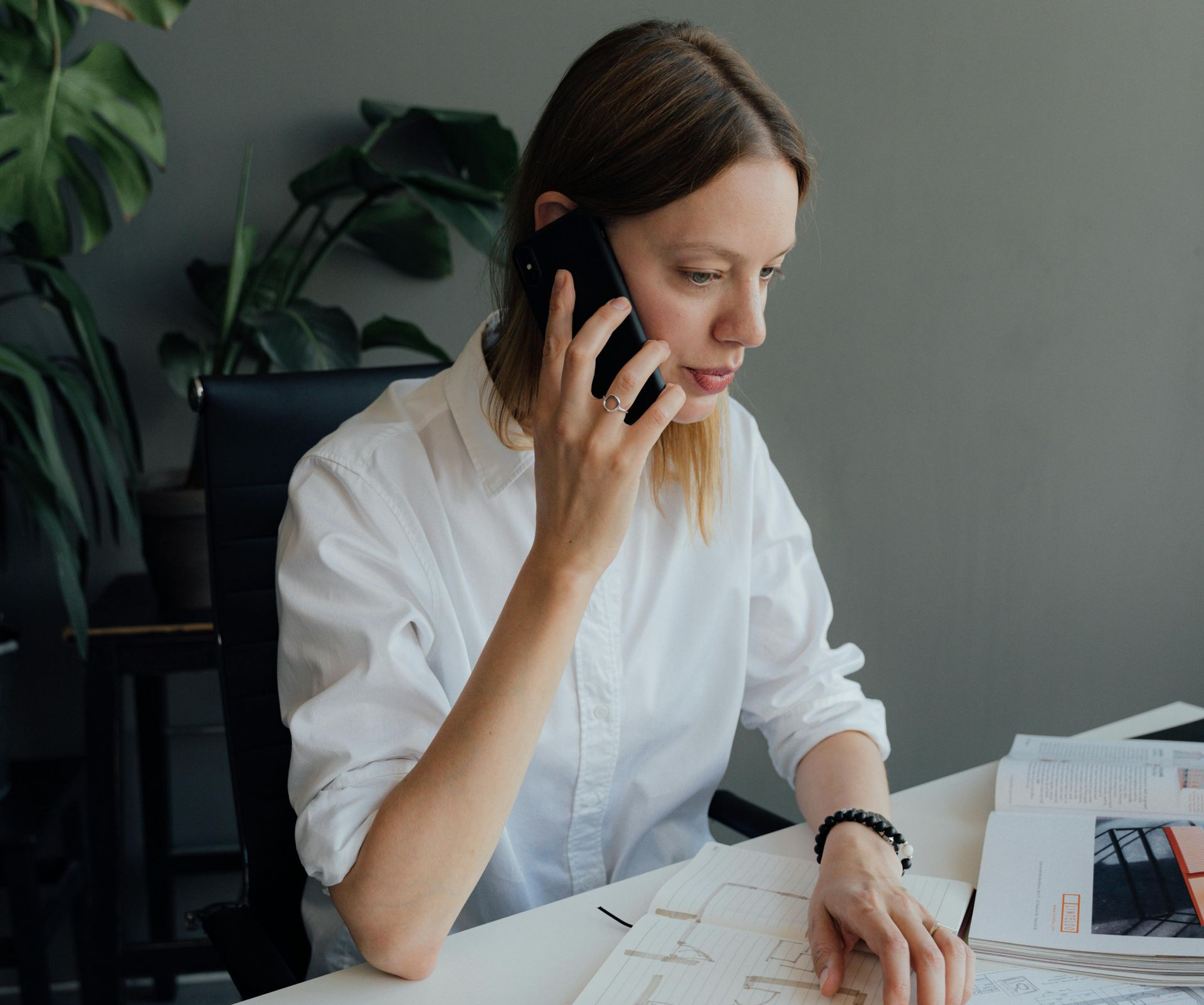 Woman in white shirt on a phone call, looking at papers at a desk, near a plant.