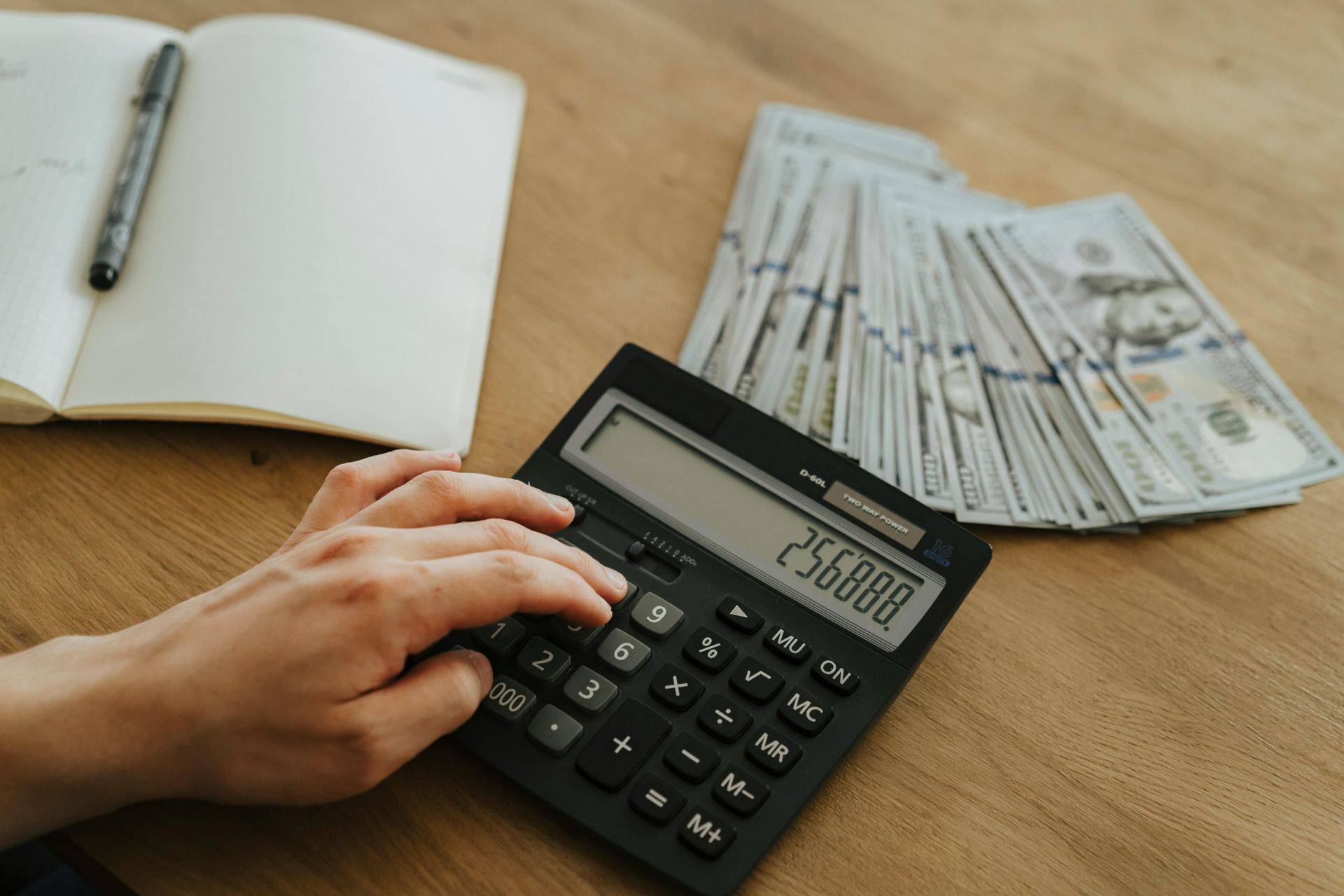 Person using a calculator with a notebook, pen, and stack of cash on a wooden surface.
