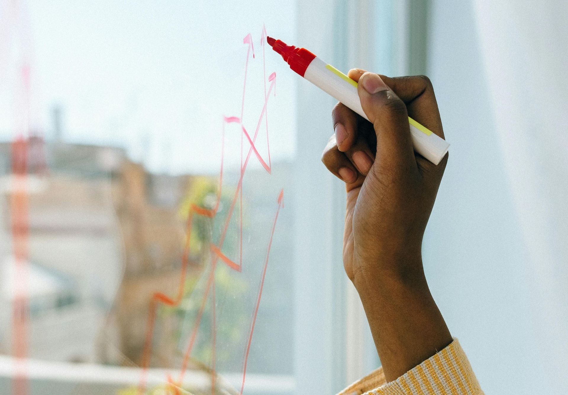 Hand holding a red marker, drawing lines on a glass surface. Building is visible outside the window.