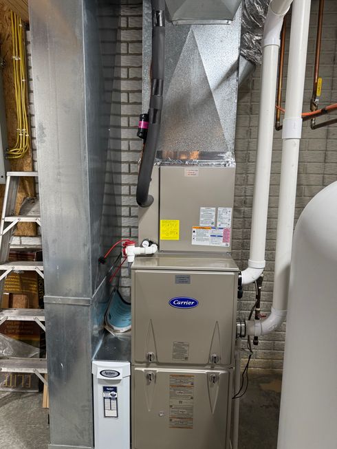 A vertical residential HVAC furnace unit installed in a basement, connected to silver ductwork and white PVC pipes.