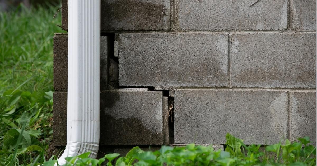 Cracked cinder block wall beside white downspout and green grass.