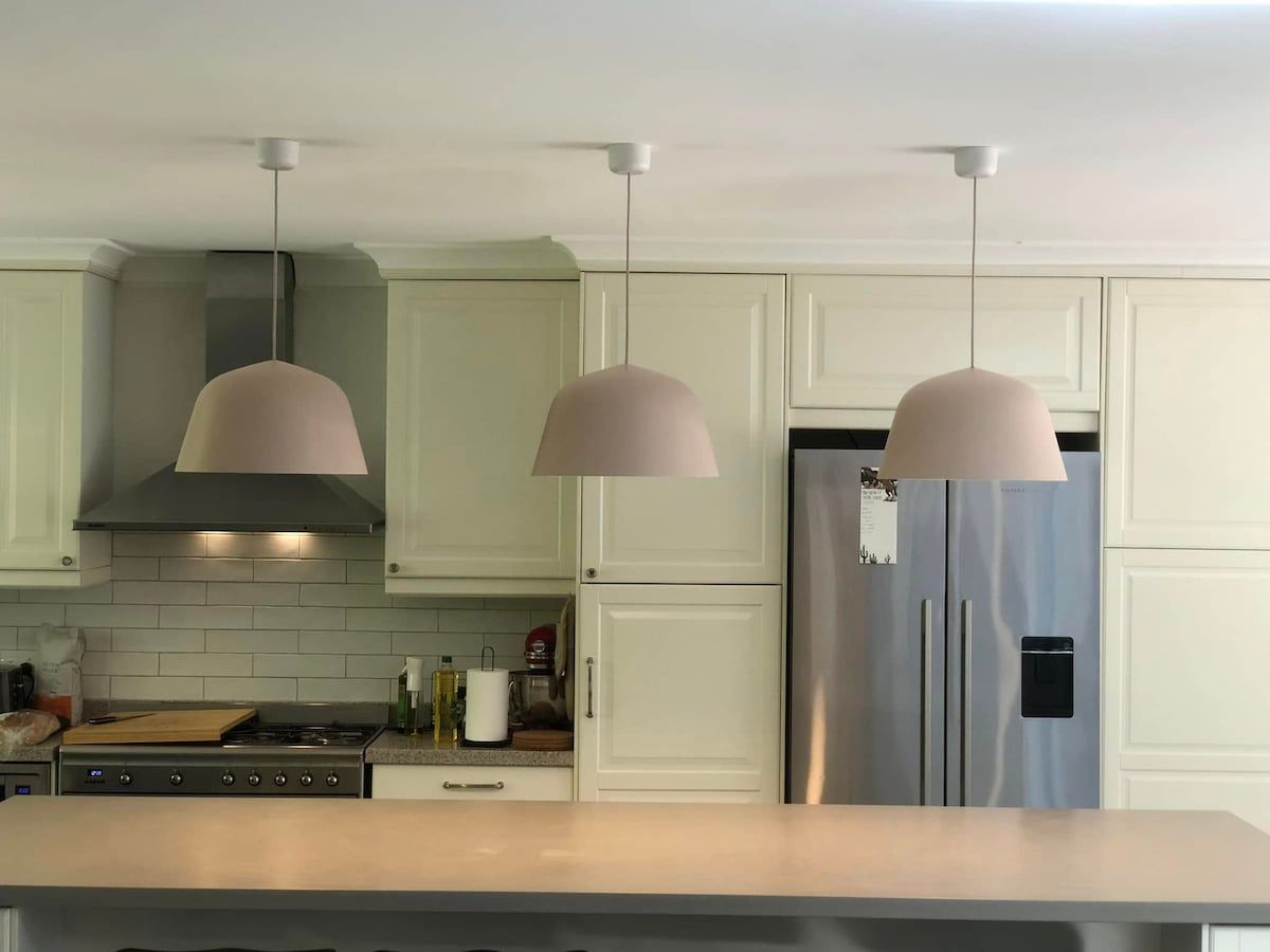 Modern kitchen with white cabinets, stainless range hood, and three pendant lights over a counter. — Technik Electrical in Bowral, NSW