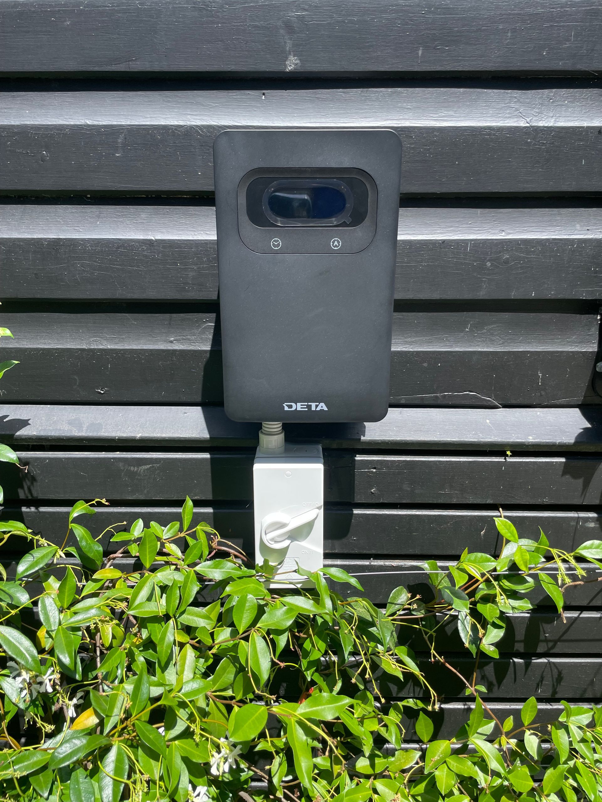 Black outdoor wall-mounted device with a camera lens above green shrubs — Technik Electrical in Renwick, NSW
