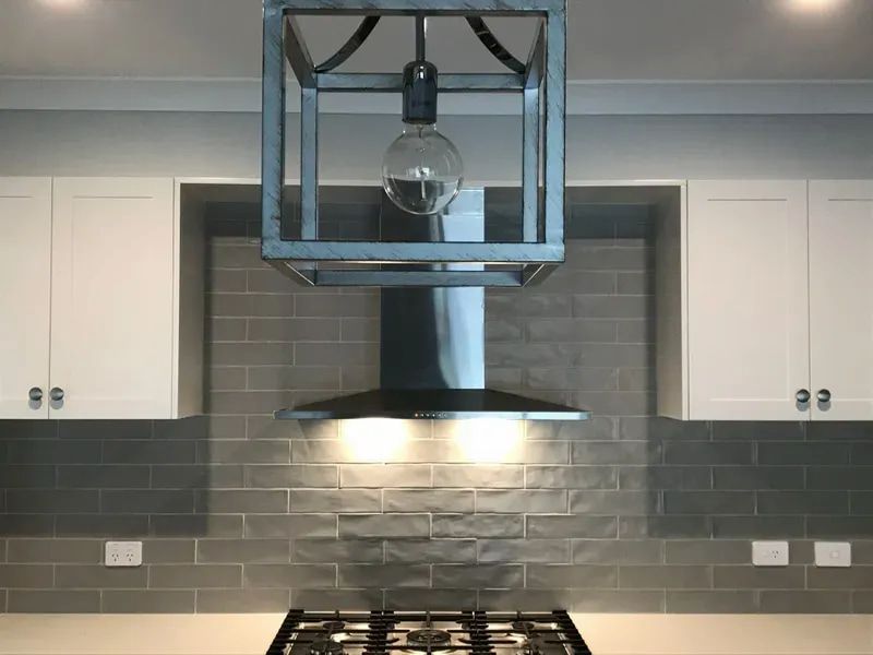 Modern kitchen with gray tile backsplash, white cabinets, range hood, and hanging light fixture — Technik Electrical in Bowral, NSW