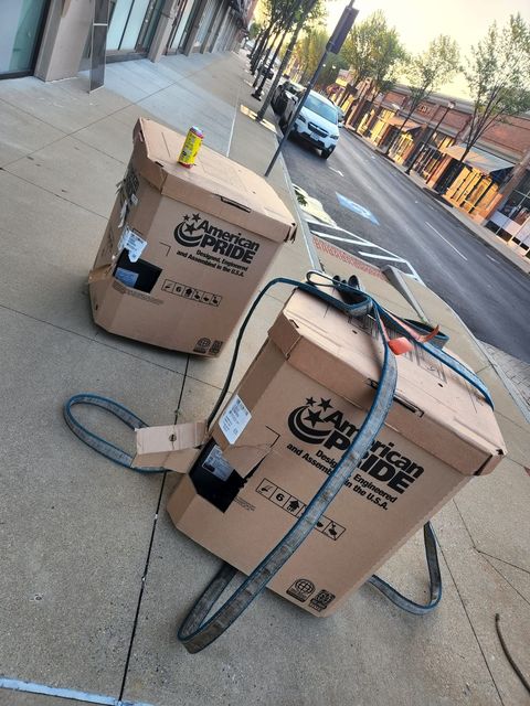 Two cardboard boxes with straps on a sidewalk. One has a yellow object on top.