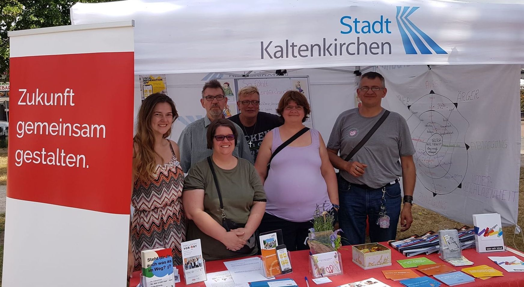 Bild von dem Projektteam beim Stadtfest. Es ist der Stand mit Flyern, Broschüren und Plakaten zu sehen und die Personen lächeln in die Kamera.