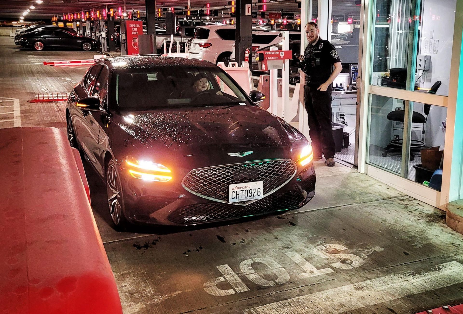 Maroon sedan at a drive-thru pickup window at night, with a worker standing beside it.