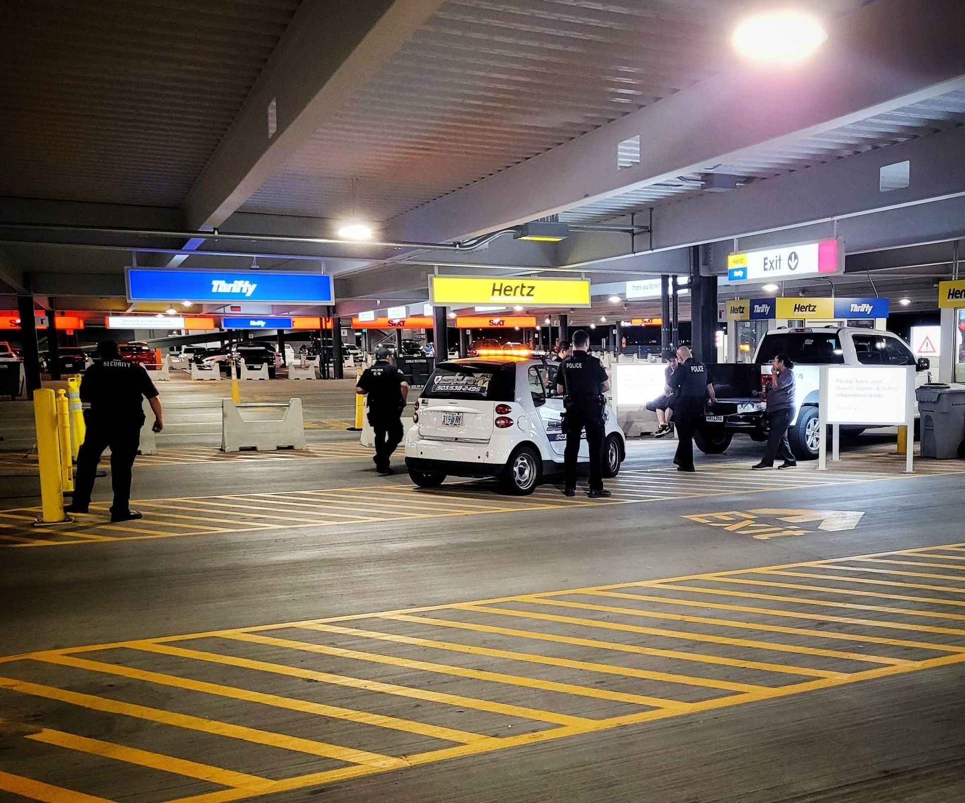 Nighttime airport curbside with people, baggage carts, and taxis under bright terminal signs.