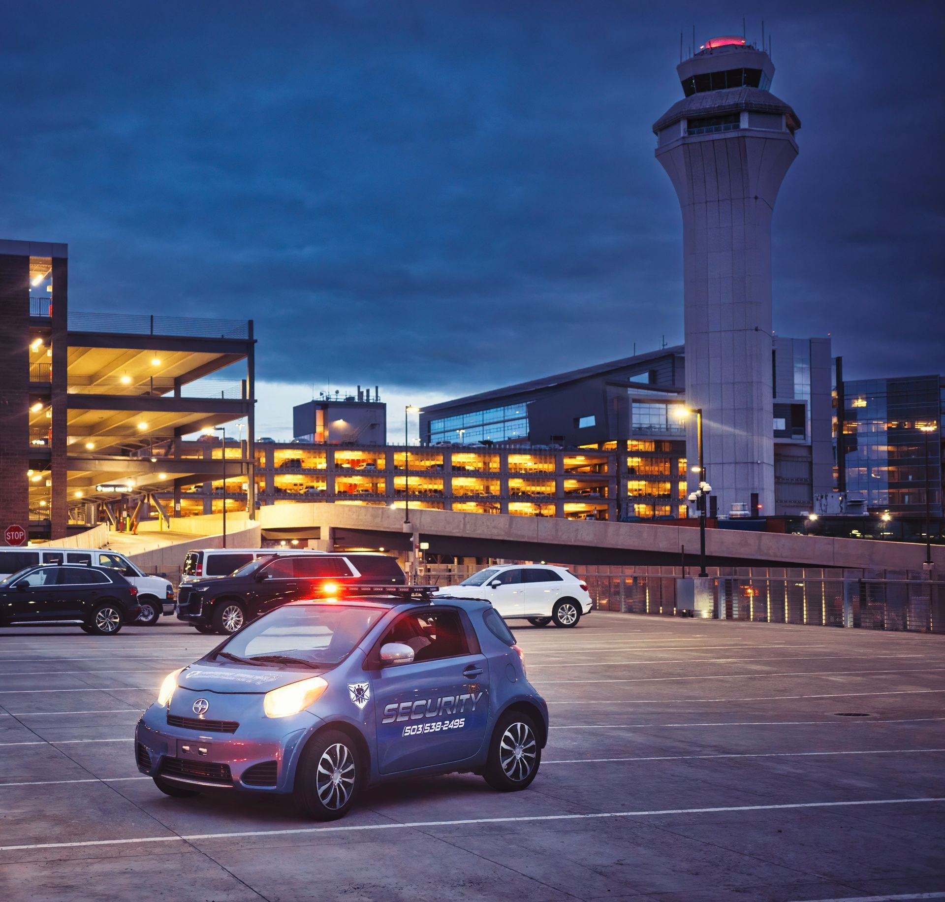 Blue Smart car parked at night outside a lit airport terminal with a control tower in the background