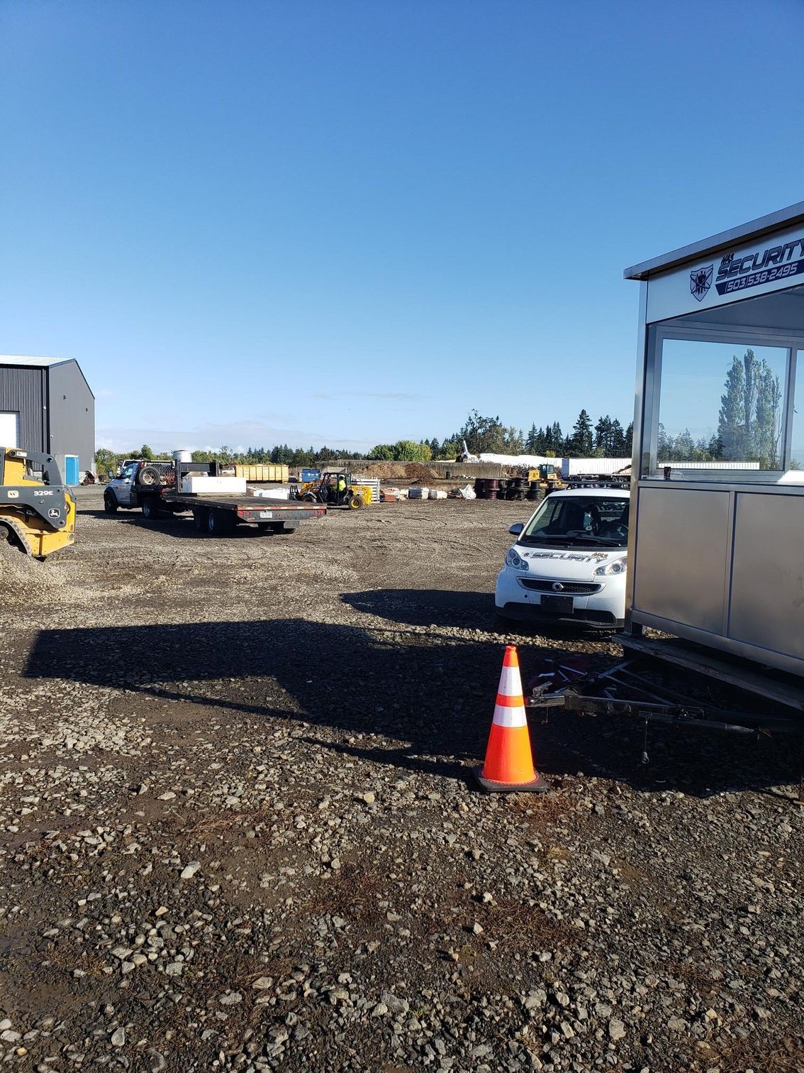 Gravel lot with equipment, a white vehicle, and an orange traffic cone beside a silver trailer